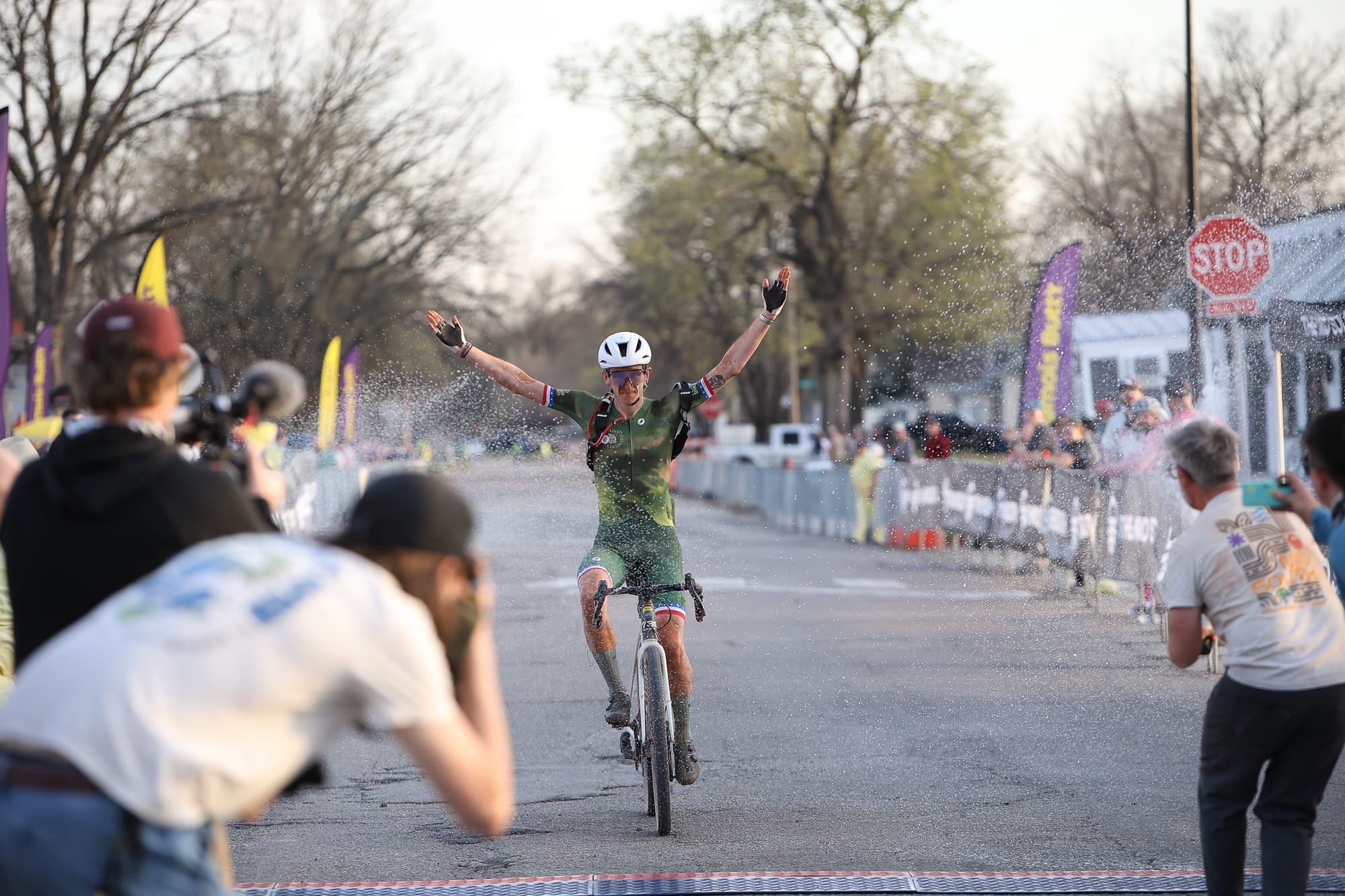 Robin Cummings raises both arms through champagne spray while crossing the finish line at the 2026 Mid South nonbinary pro race Friday at Block 34 in Stillwater, Oklahoma.