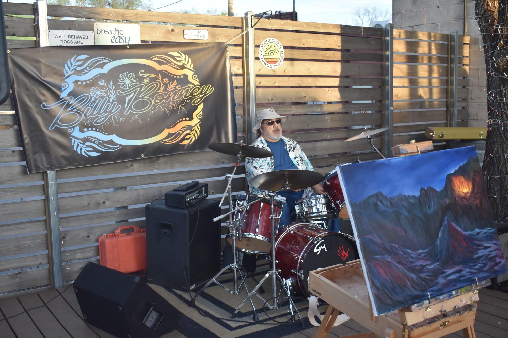 Drummer Greg Miller plays behind a Billy Bonney banner at Stonecloud Brewing, with the hand-painted As The Canyons Roar album cover on an easel to his right. 