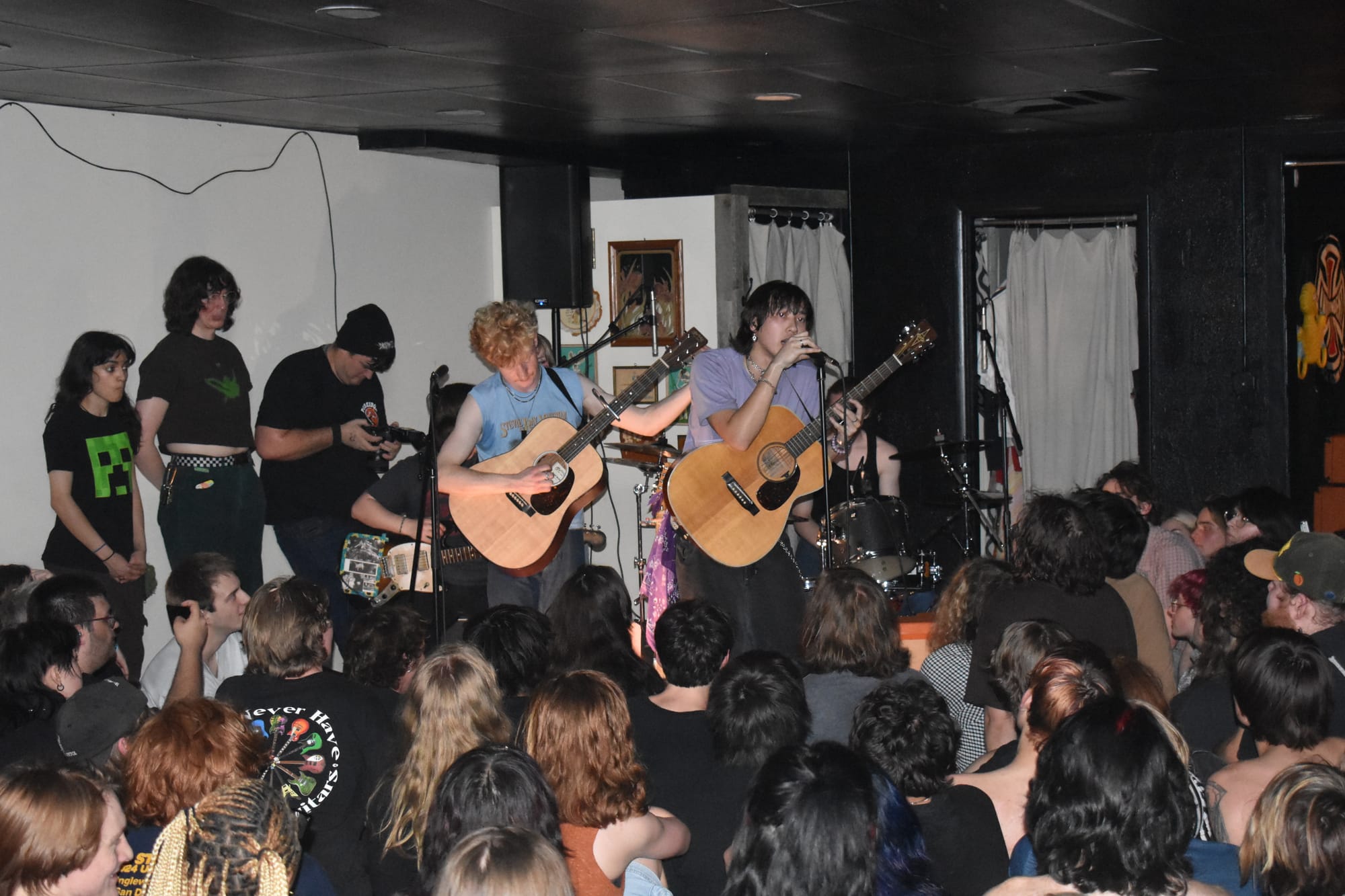 Two acoustic guitarists perform on a low stage at a small indoor venue while a large crowd sits on the floor in front of them.