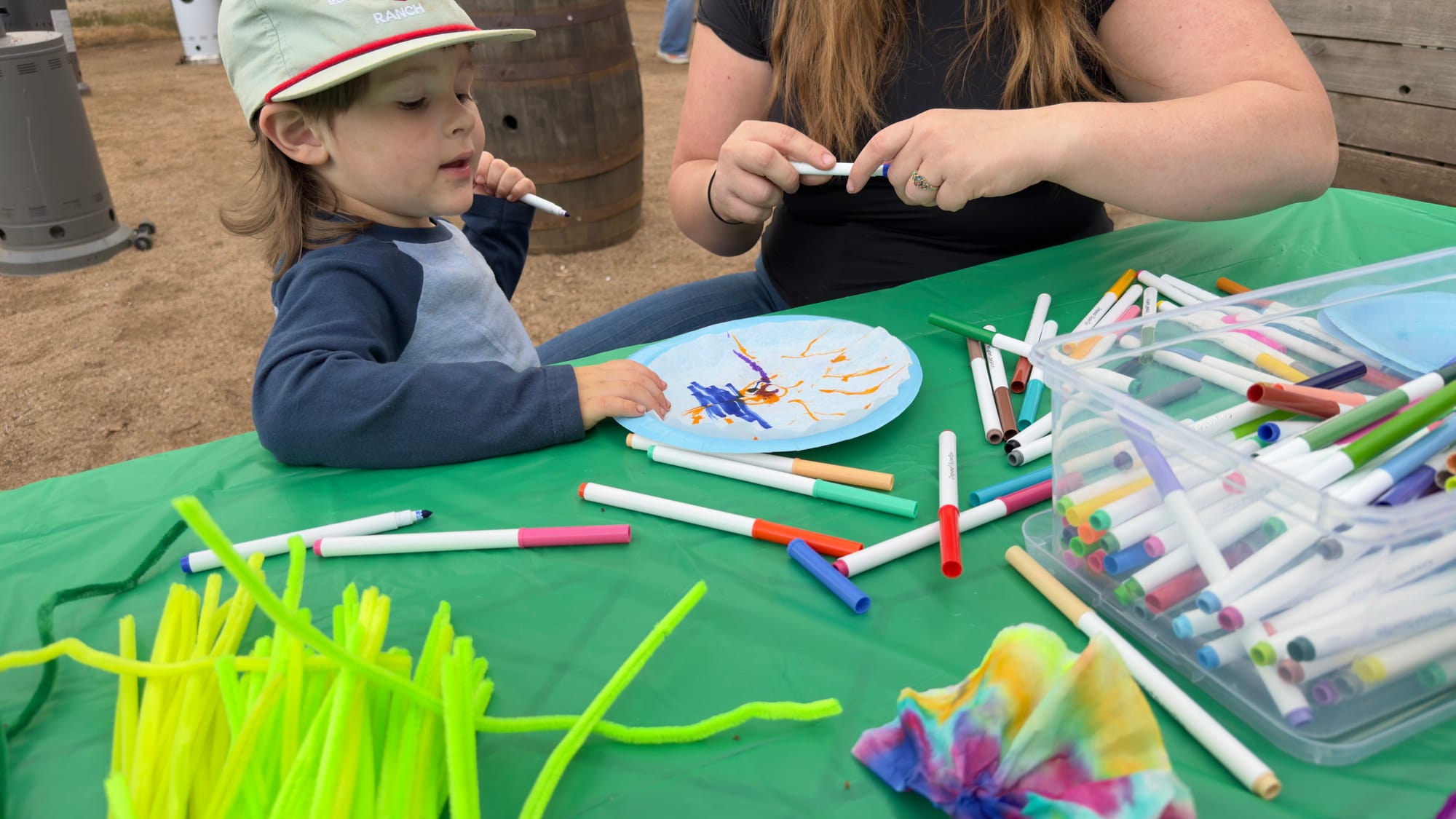 A young child studies a colorful paper plate at a green craft table scattered with markers and pipe cleaners during a StillWonder event at Stonecloud Patio & Taproom in Stillwater.