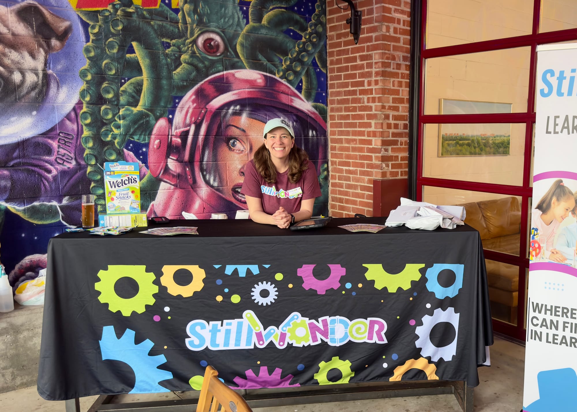 Sara Brown sits smiling behind a StillWonder-branded table at Stonecloud Patio & Taproom in Stillwater, with a large sci-fi mural on the brick wall behind her.
