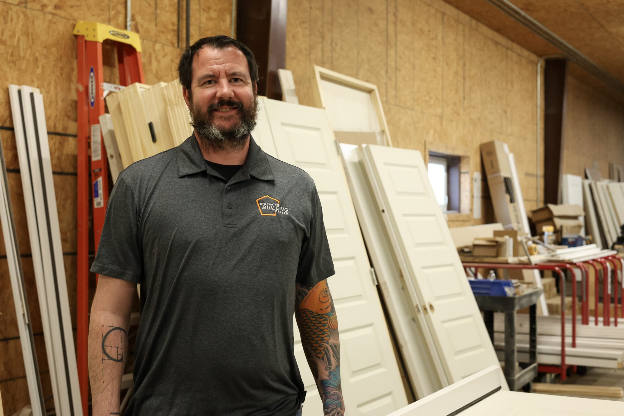 James Morrison, Director of Operations at Stillwater Building Center, stands smiling in the door shop surrounded by finished door panels leaning against the wall.