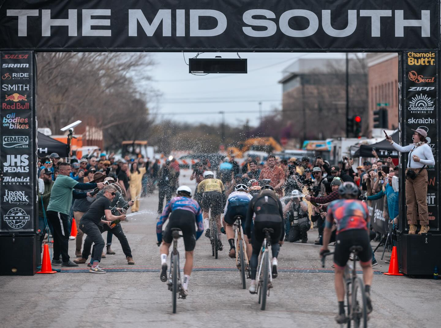 A five-rider sprint group crosses the 2024 Mid South finish line arch as champagne sprays from both sides of the chute in front of a packed crowd on Husband Street.