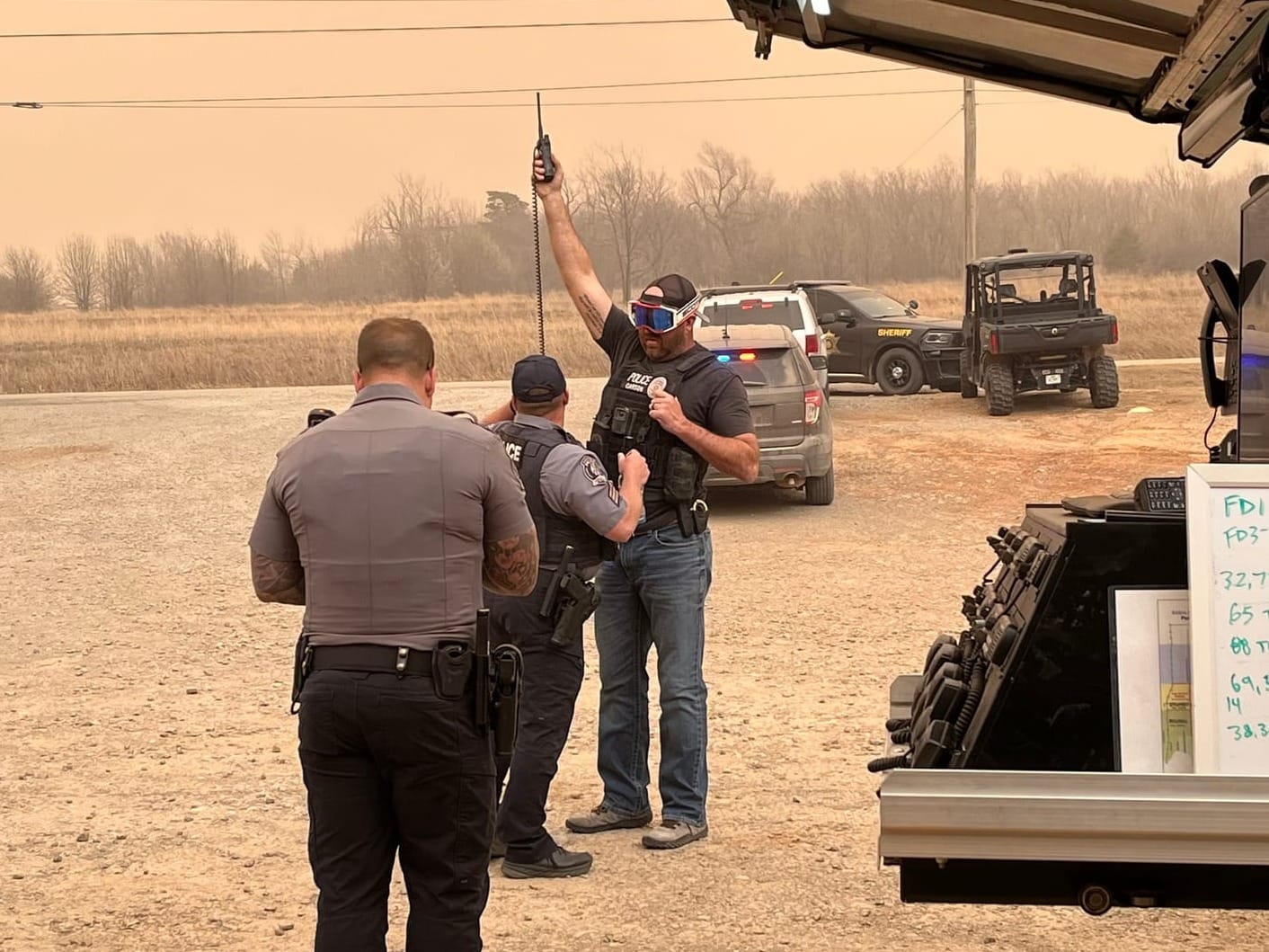A police officer holds a radio overhead at an outdoor command post as smoke fills the sky behind him during the March 14, 2025 Stillwater wildfires.