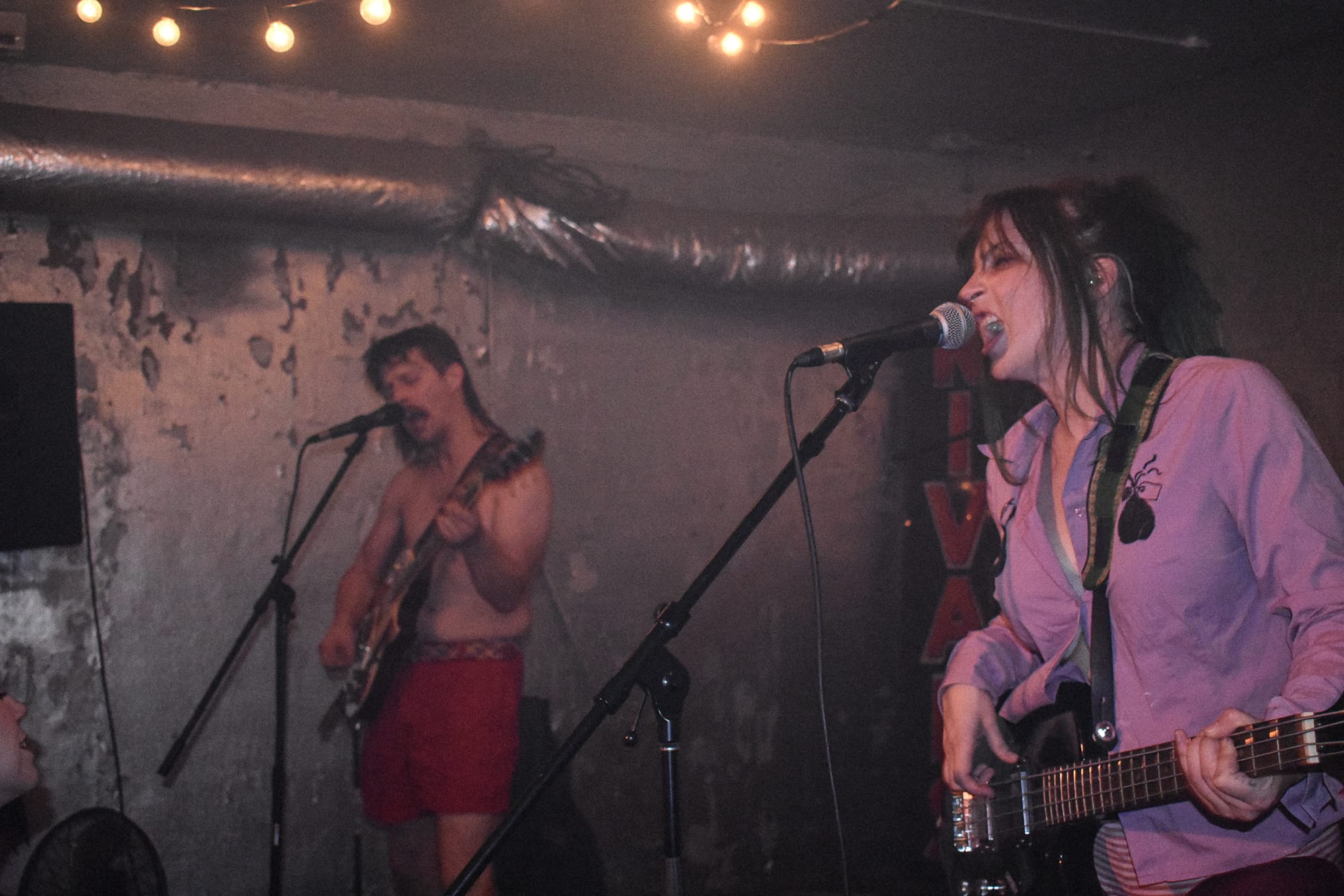 Ugly Cowboys bassist Bee McAdoo sings into a microphone while guitarist Sam Miess performs shirtless behind her on stage at Velvet Fudge.