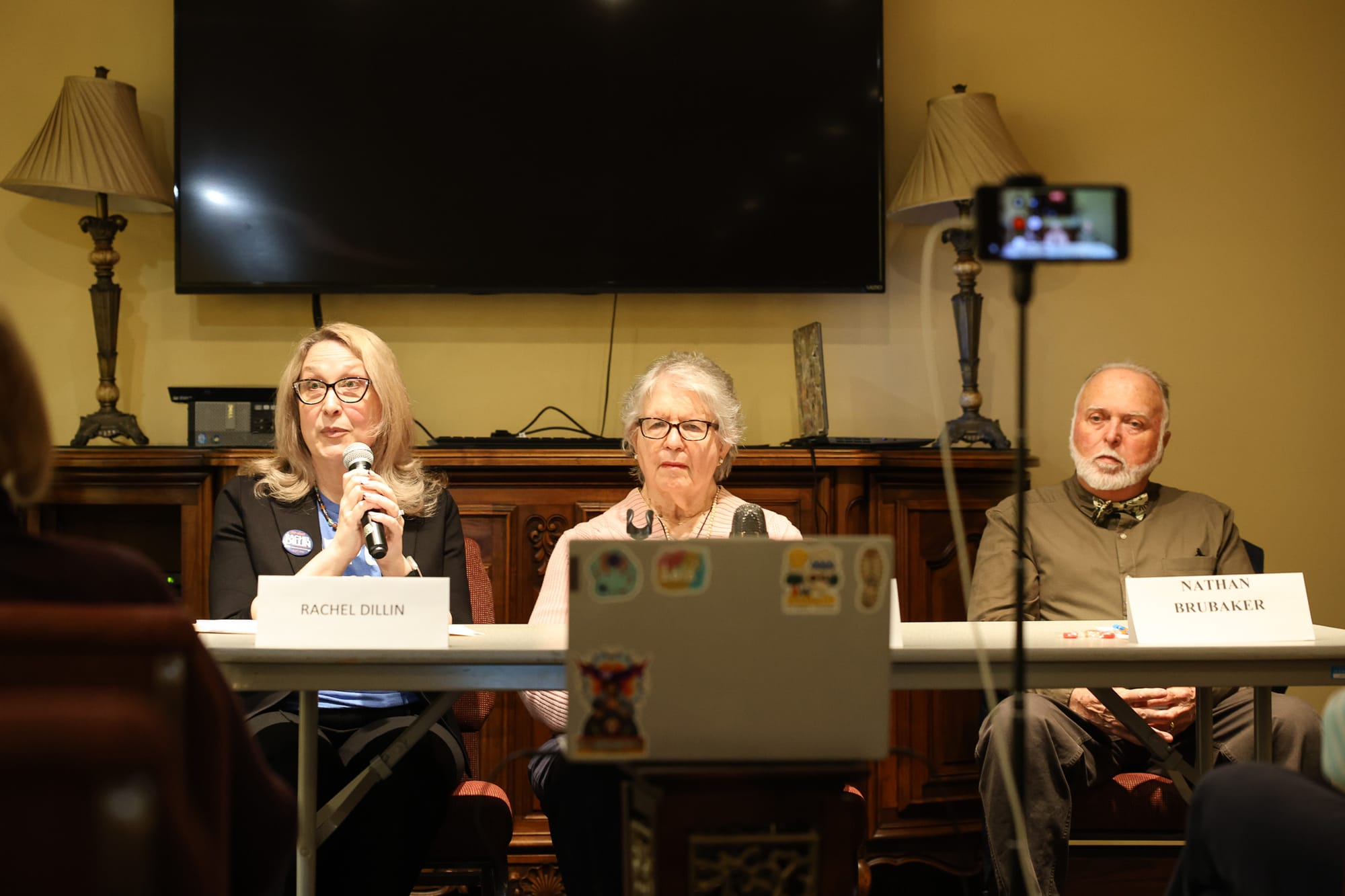 Rachel Dillin speaks into a microphone at a candidate forum table, flanked by Joanne Murer in the center and Nathan Brubaker on the right. Nameplates for Dillin and Brubaker are visible on the table.