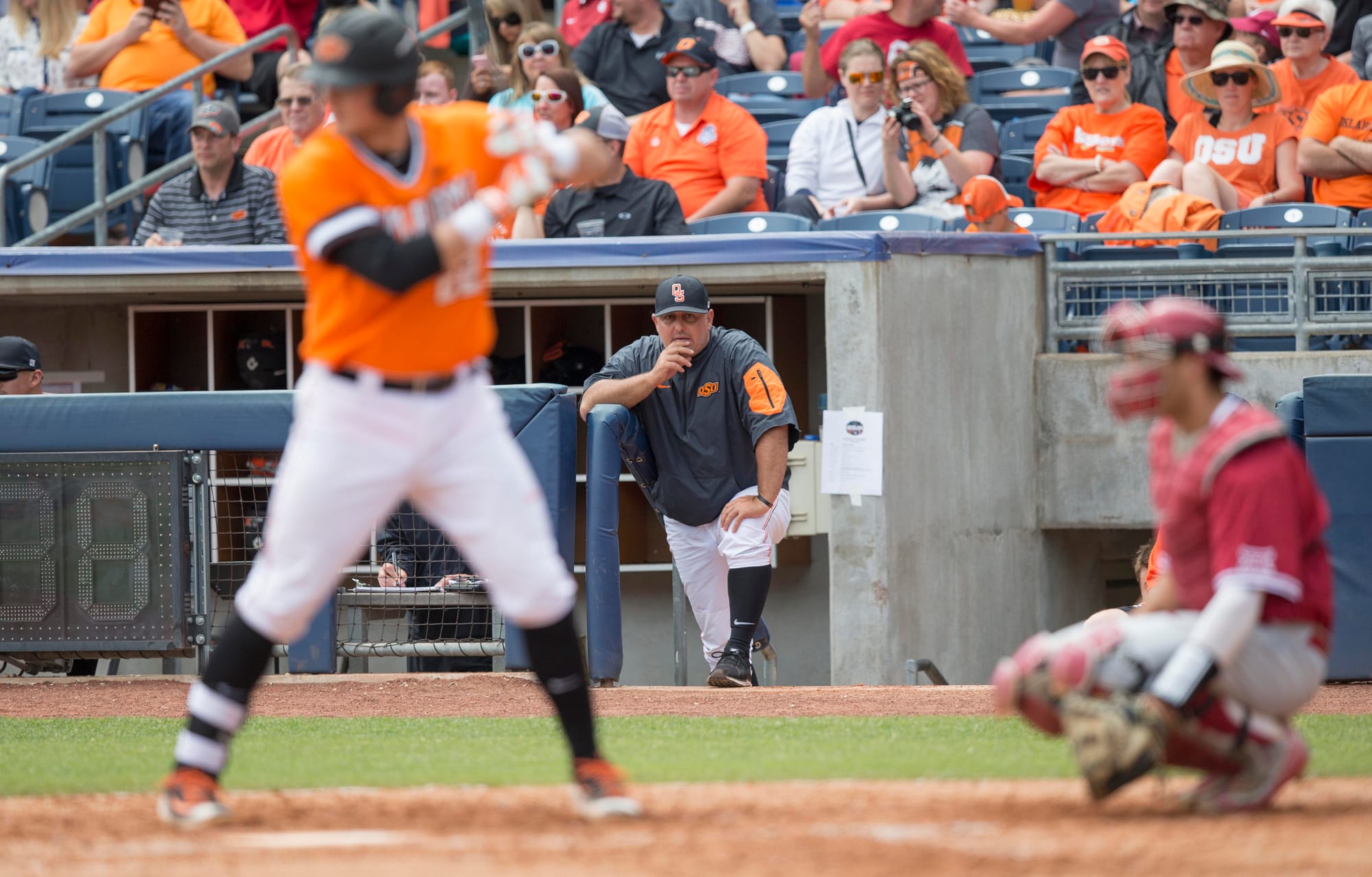 OSU head coach Josh Holliday leans forward from the dugout steps, watching intently during a 2016 Bedlam baseball game against Oklahoma with an OSU batter at the plate.