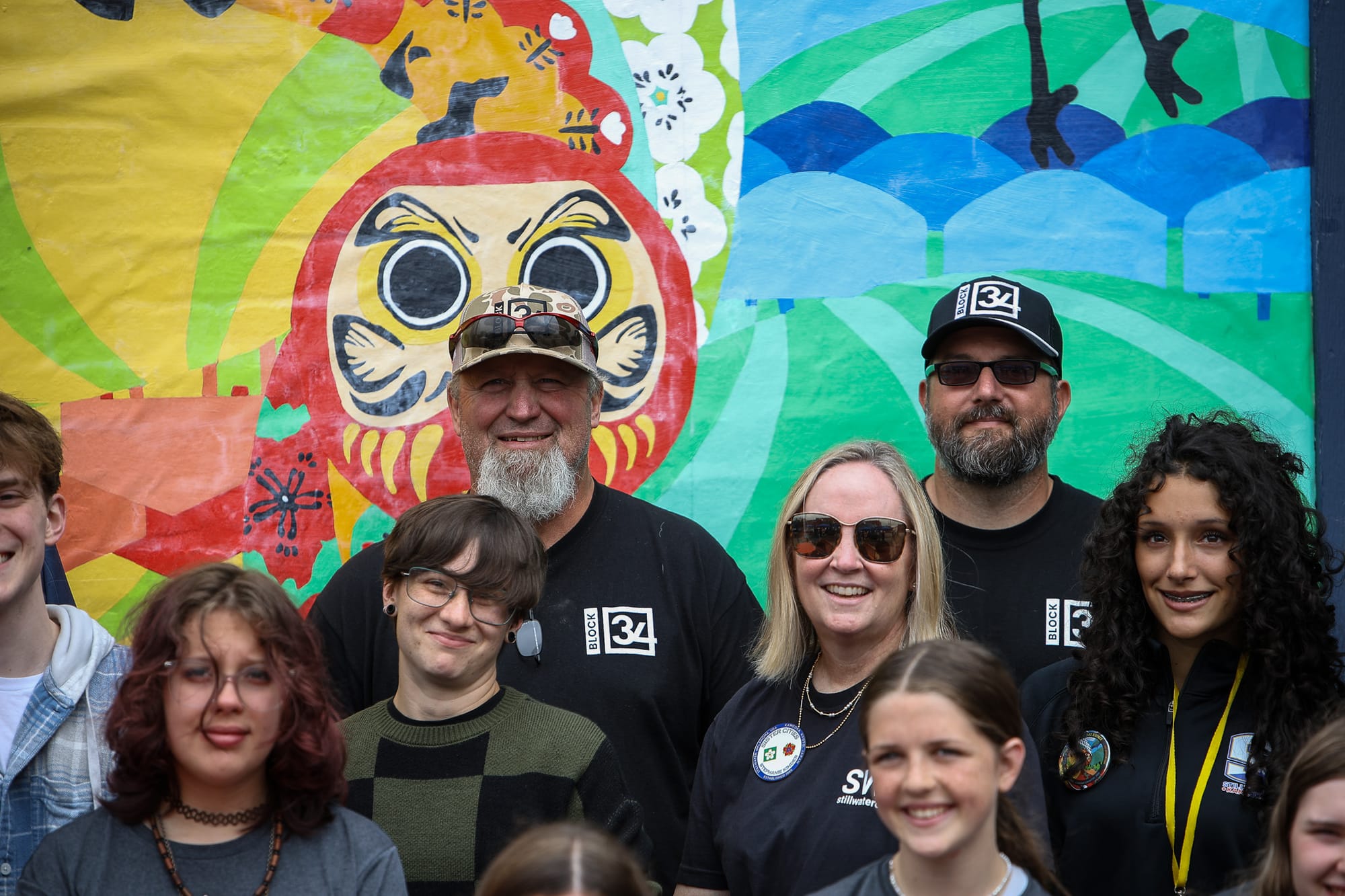 Monty Ridgway and Andy Woods, wearing Block 34 shirts, stand behind Bec Summer and Stephanie Kinder in front of a colorful mural featuring a large daruma doll at Chris Salmon Plaza.