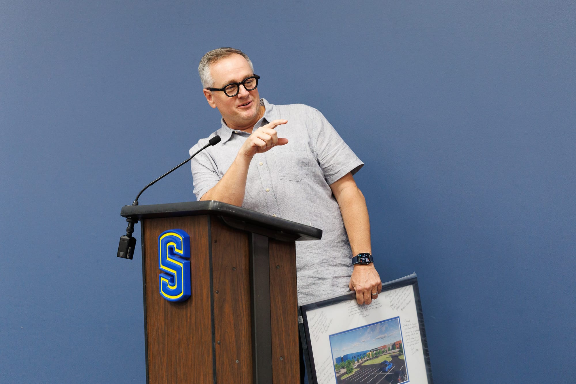 Dr. Marshall Baker speaks at a podium during his recognition at the June 23, 2026, Stillwater Board of Education meeting, holding a framed aerial photo of the new high school signed by board members.