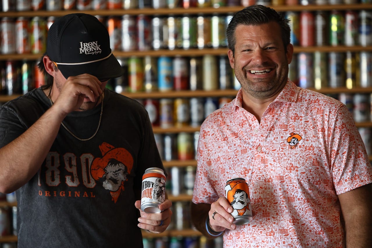 Trenton Inselman, left, adjusts his cap while holding a can of Cowboy Blonde, and Brady Sidwell laughs while holding a can of Original 1890, in front of a wall of beer cans.
