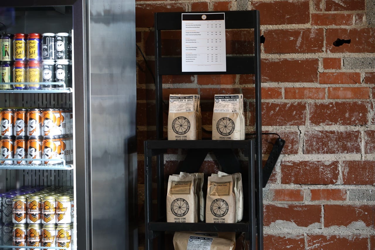 Bags of Chisholm Trail Milling flour, including Gallagher and Butler's Gold wheat varieties, sit on a shelf next to a cooler of Iron Monk beer cans in the taproom.