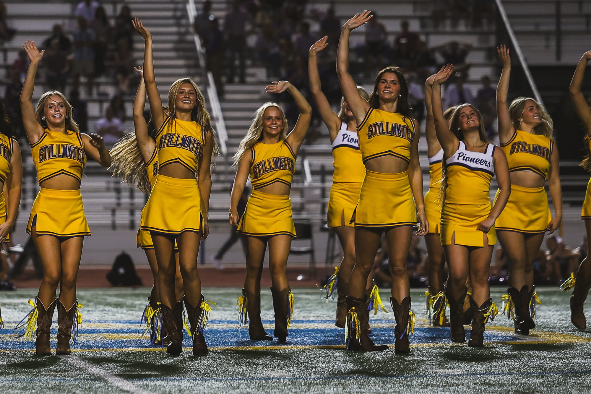 Stillwater pom squad members in gold uniforms and cowboy boots wave to the crowd on the field at Pioneer Stadium, with senior Daisy Goff at center.