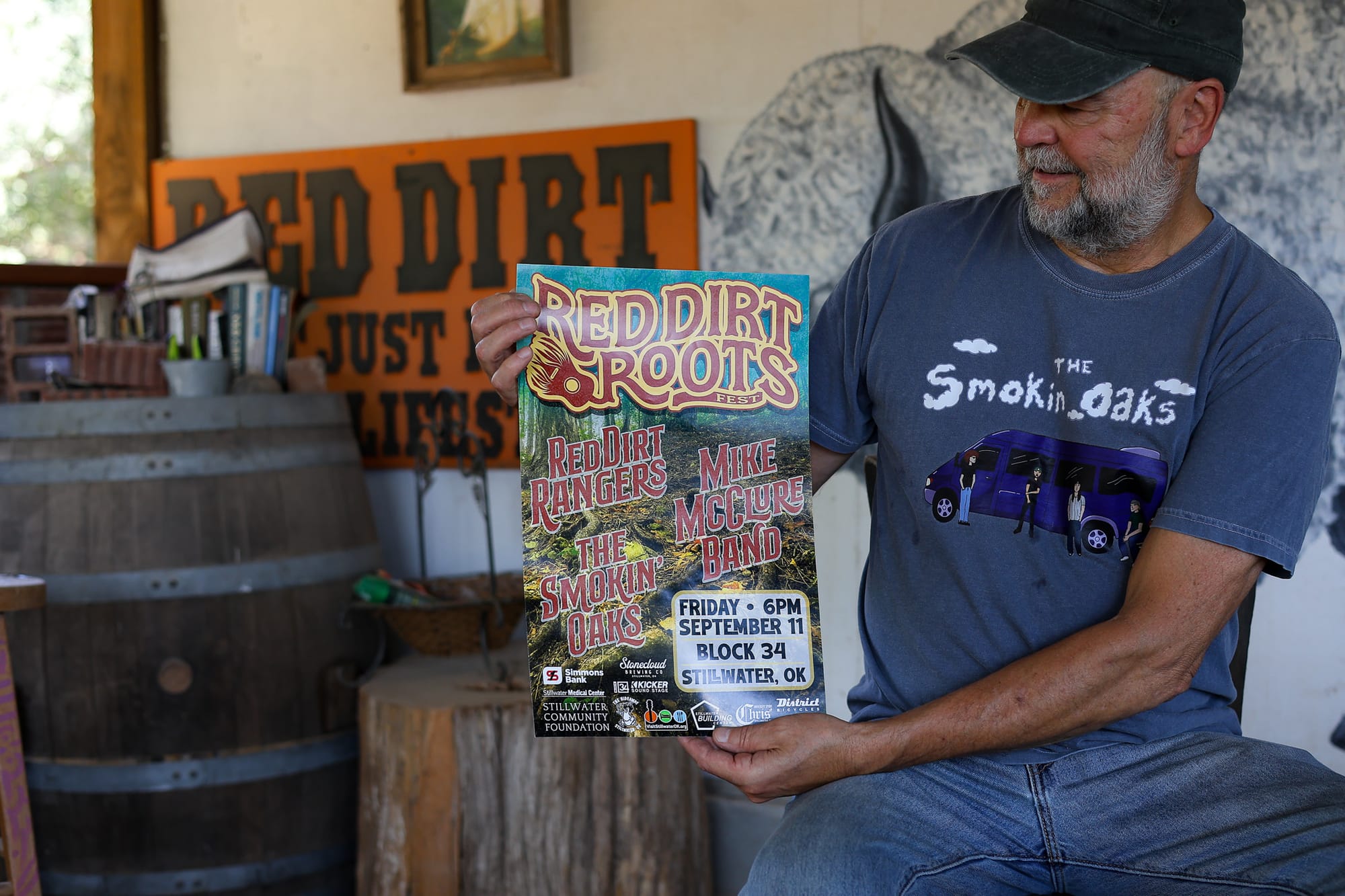 John Cooper of the Red Dirt Rangers holds a poster for Red Dirt Roots Fest while seated at The Farm near Stillwater, Oklahoma, wearing a Smokin' Oaks T-shirt and cap.