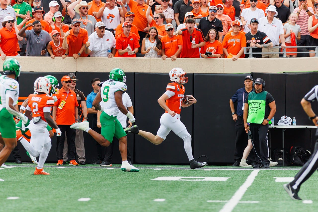 Oklahoma State quarterback Drew Mestemaker in an orange jersey sprints downfield along the sideline with the football as an Oregon defender pursues him at Boone Pickens Stadium.