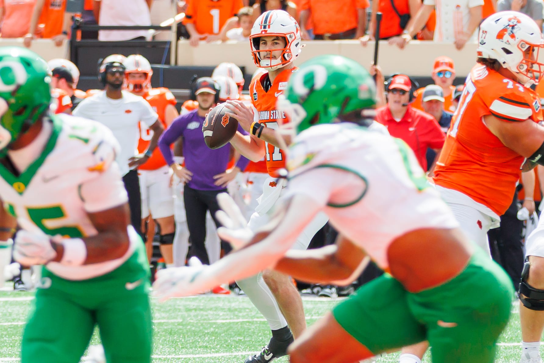 Oklahoma State quarterback Drew Mestemaker in an orange jersey sets to throw as Oregon defenders in white and green rush toward him at Boone Pickens Stadium.