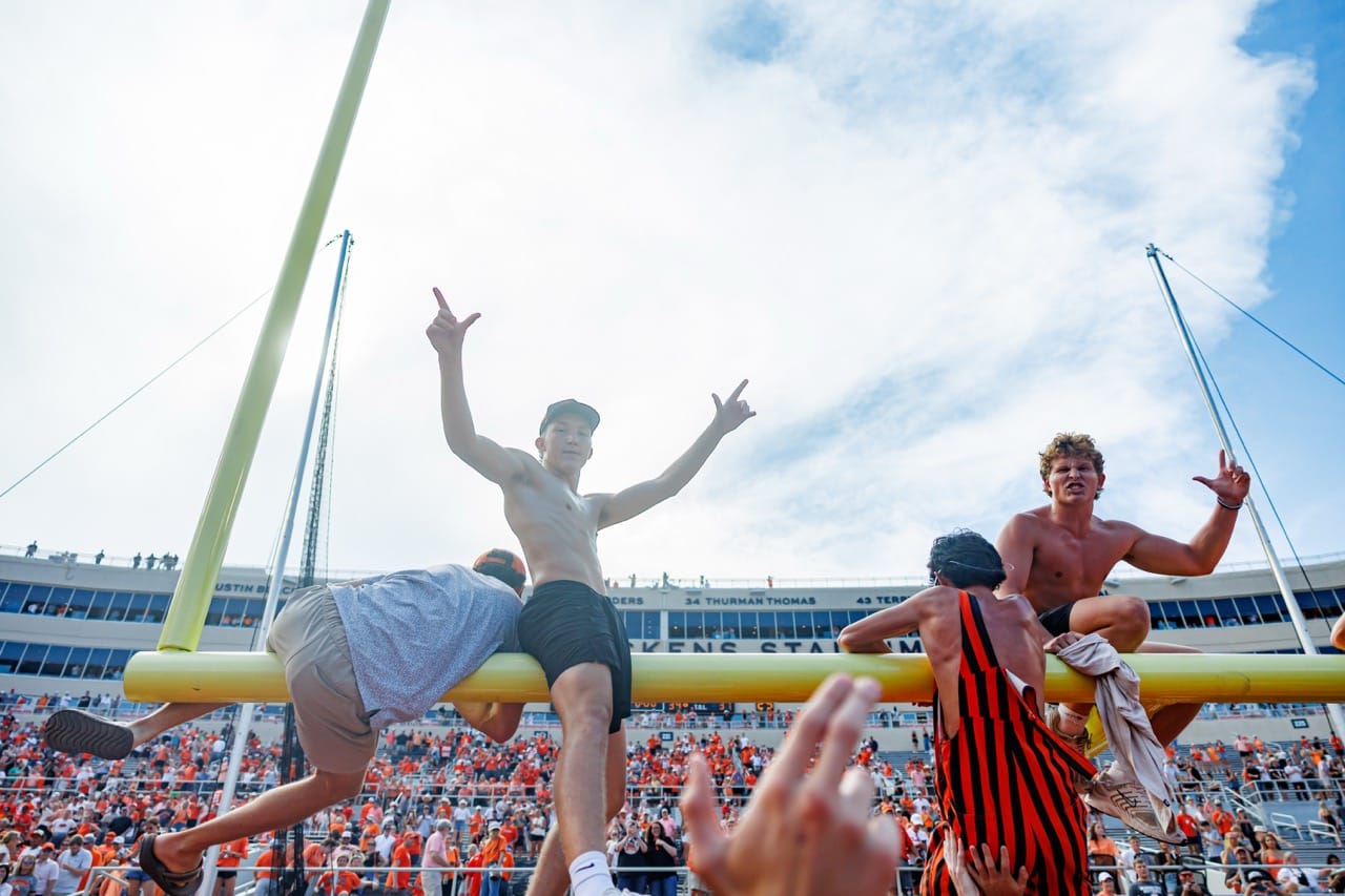 Shirtless Oklahoma State fans sit and climb on a yellow goal post crossbar with arms raised in celebration as orange-clad crowds fill the stands at Boone Pickens Stadium.