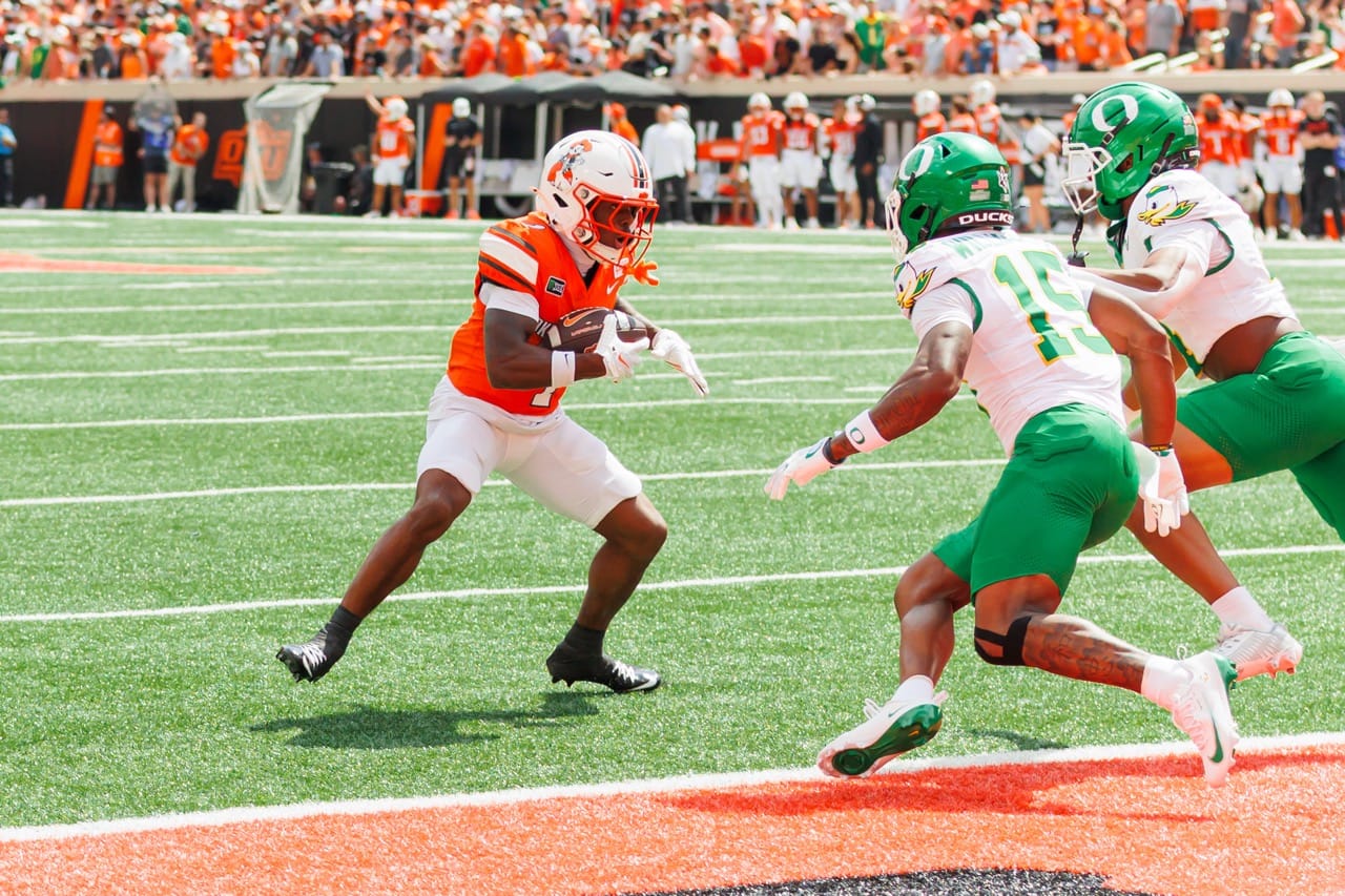 Oklahoma State receiver Miles Coleman in an orange jersey cuts upfield with the football as two Oregon defenders in white and green close in near the goal line at Boone Pickens Stadium.