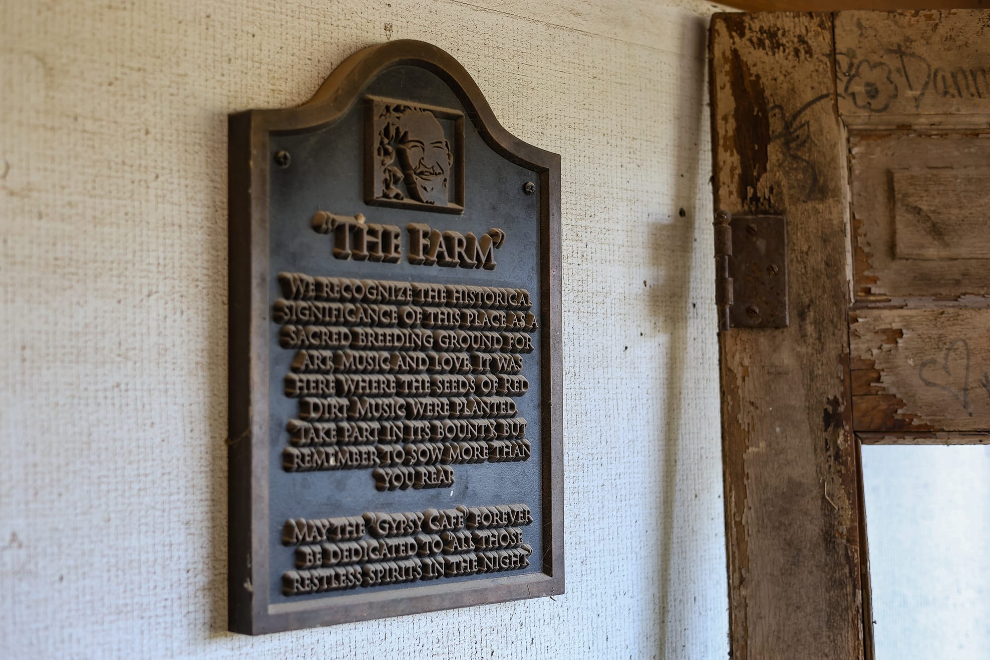 A weathered plaque titled "The Farm" hangs on an interior wall near a wooden door frame, bearing a relief portrait and text dedicating the property to Red Dirt music's origins and the Gypsy Cafe.