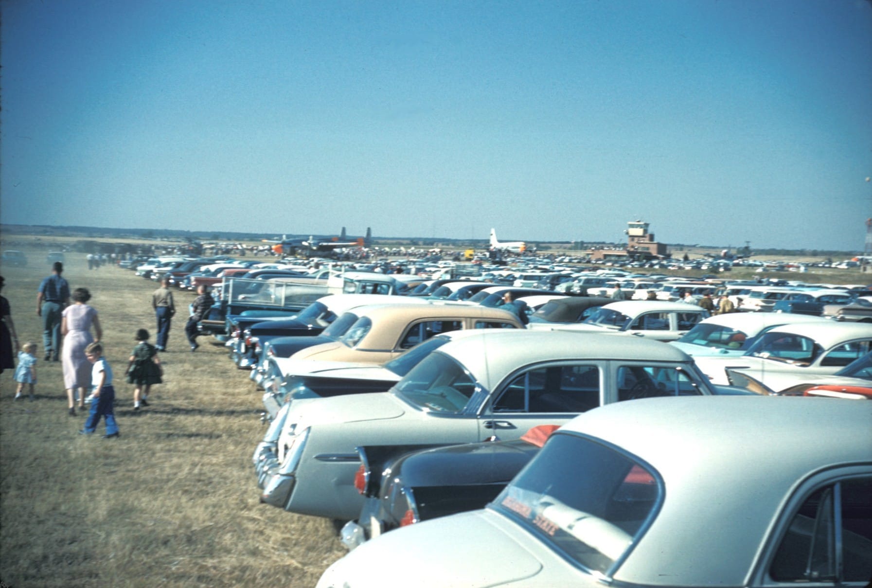A woman in a pink dress walks with young children beside rows of 1950s cars parked on grass, with military planes, a small terminal and a control tower on the horizon in 1960.