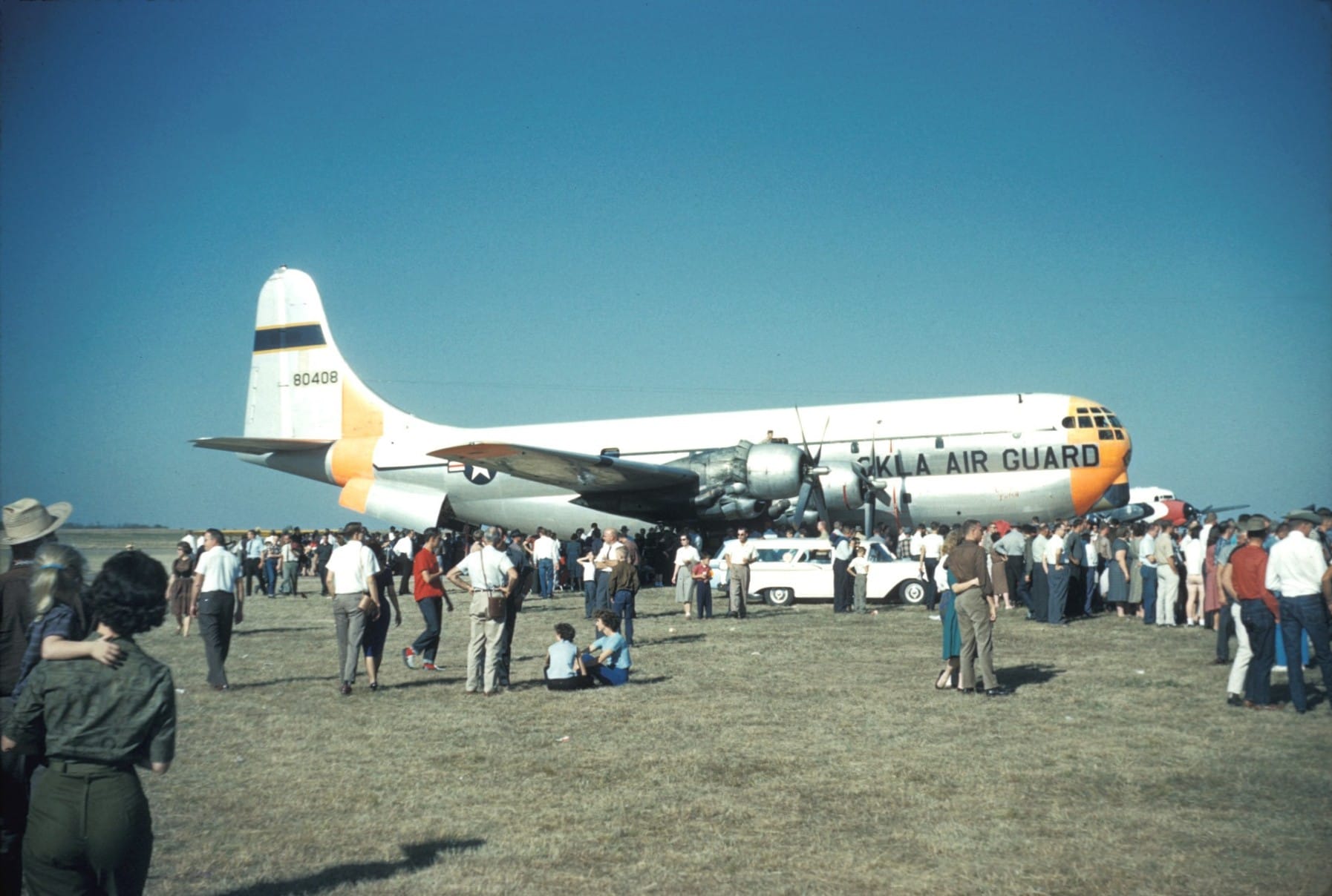 Crowds gather on the grass around a large silver and white four-engine cargo plane marked OKLA AIR GUARD, with orange nose and tail trim, at an airport in 1960.