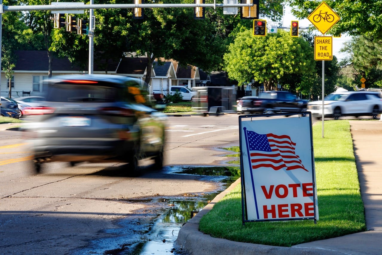 A "Vote Here" sign with an American flag stands on a curb as a car blurs past at an intersection in Stillwater on primary election day, Tuesday, June 16, 2026.