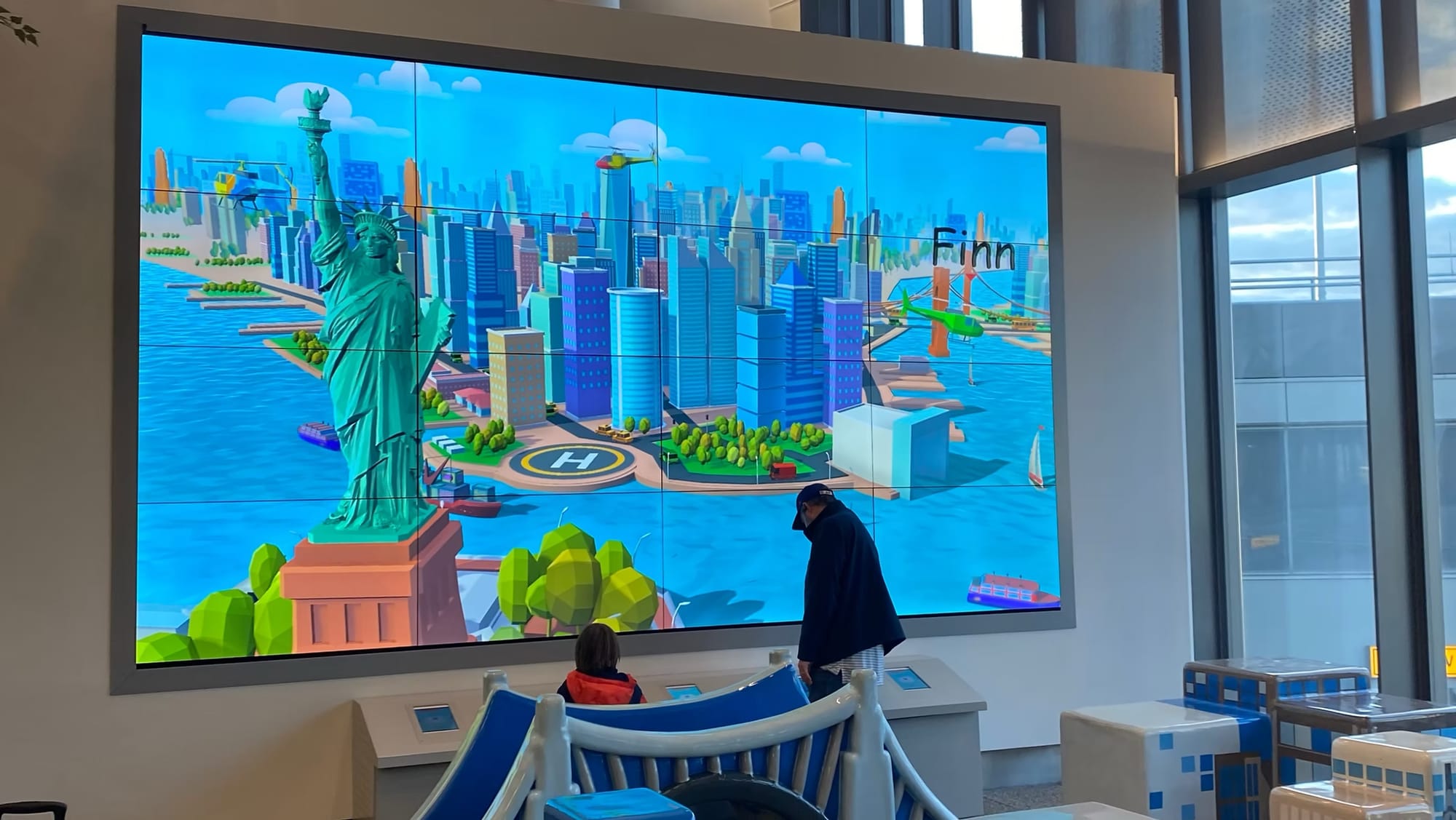 A child and an adult stand at scanning stations below a 16-screen video wall showing an animated New York City scene with the Statue of Liberty, helicopters and boats.
