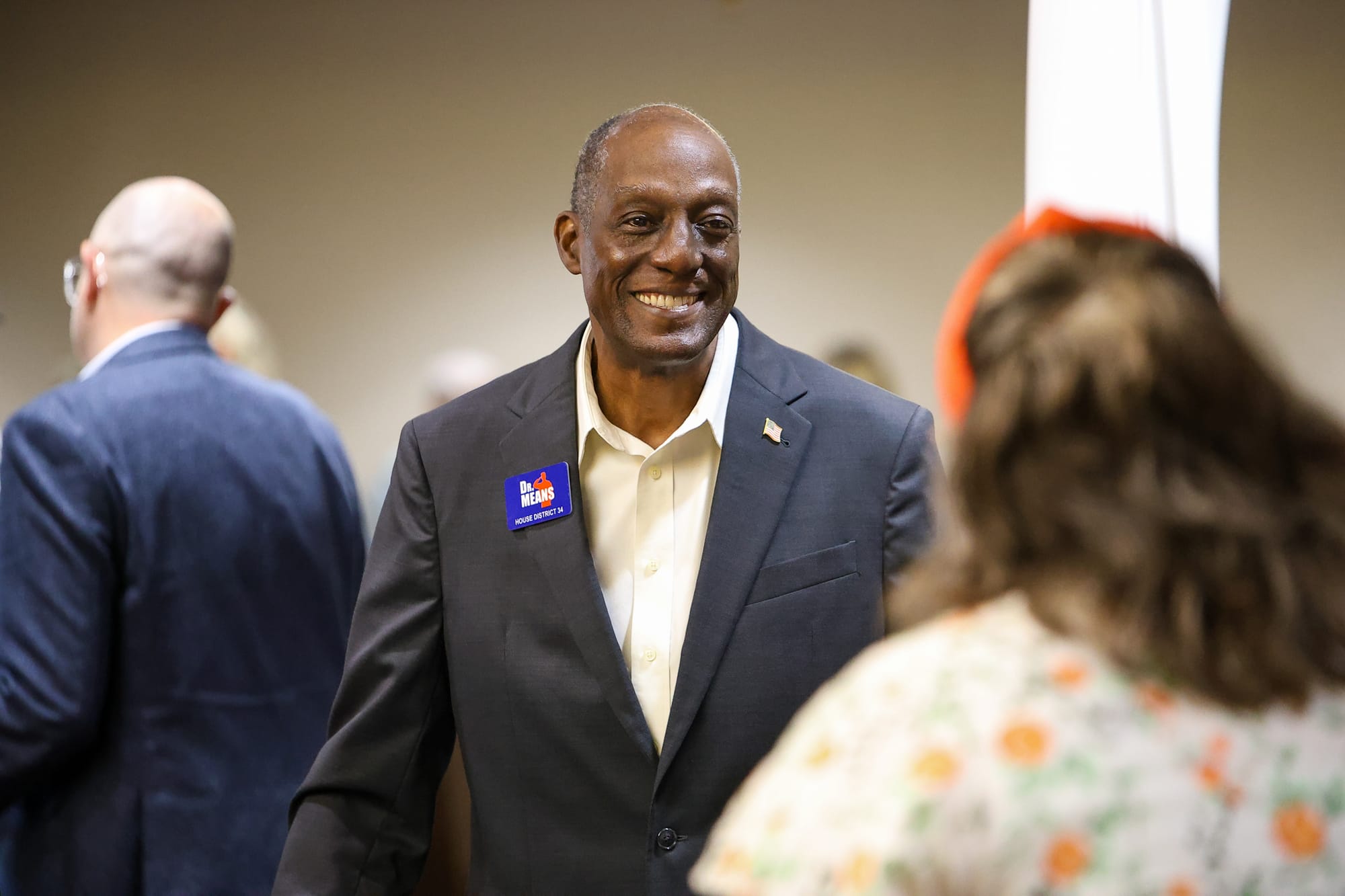 Aaron Means, wearing a dark blazer and a Dr. Means House District 34 campaign badge, smiles as he greets an attendee at a Stillwater Rotary Club forum on Oct. 22, 2026.