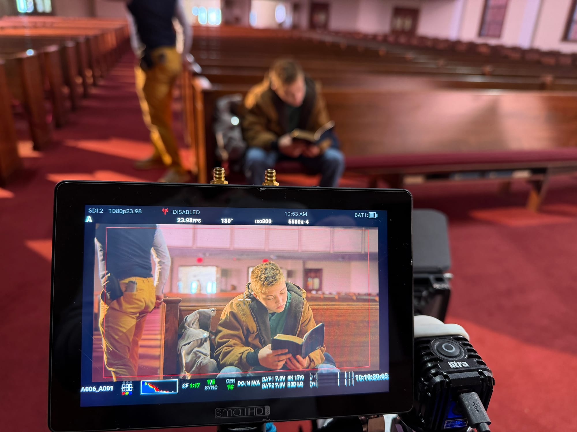 A camera monitor in the foreground shows a teen in a tan canvas jacket reading a small book while seated in a wooden church pew. Behind the monitor, the same scene is out of focus in a sanctuary with red carpet and rows of pews, and a crew member stands in the aisle.