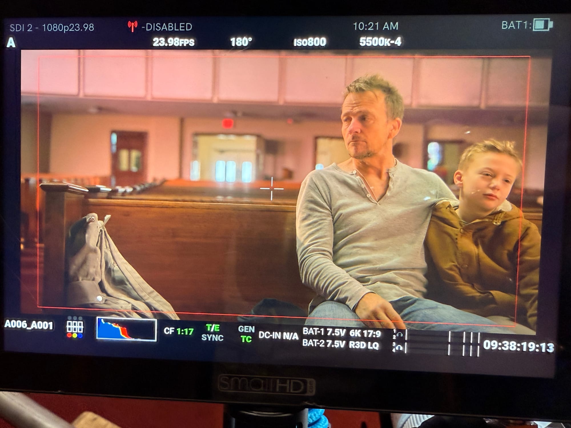 A camera monitor displays a man in a gray henley and a young boy in a tan hooded jacket sitting together in a wooden church pew, the man looking off to the side with a somber expression.
