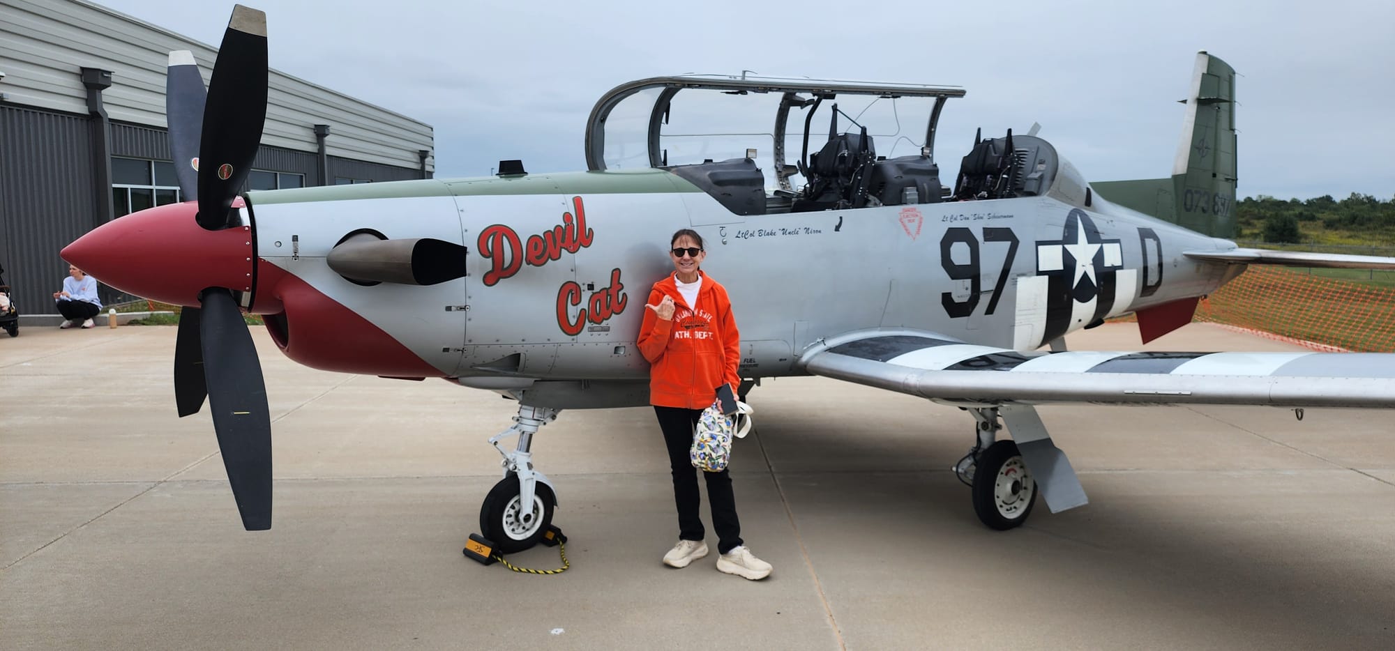 A smiling woman in an orange Oklahoma State hoodie and sunglasses points to a silver propeller plane with Devil Cat painted on its nose and World War II-style markings.