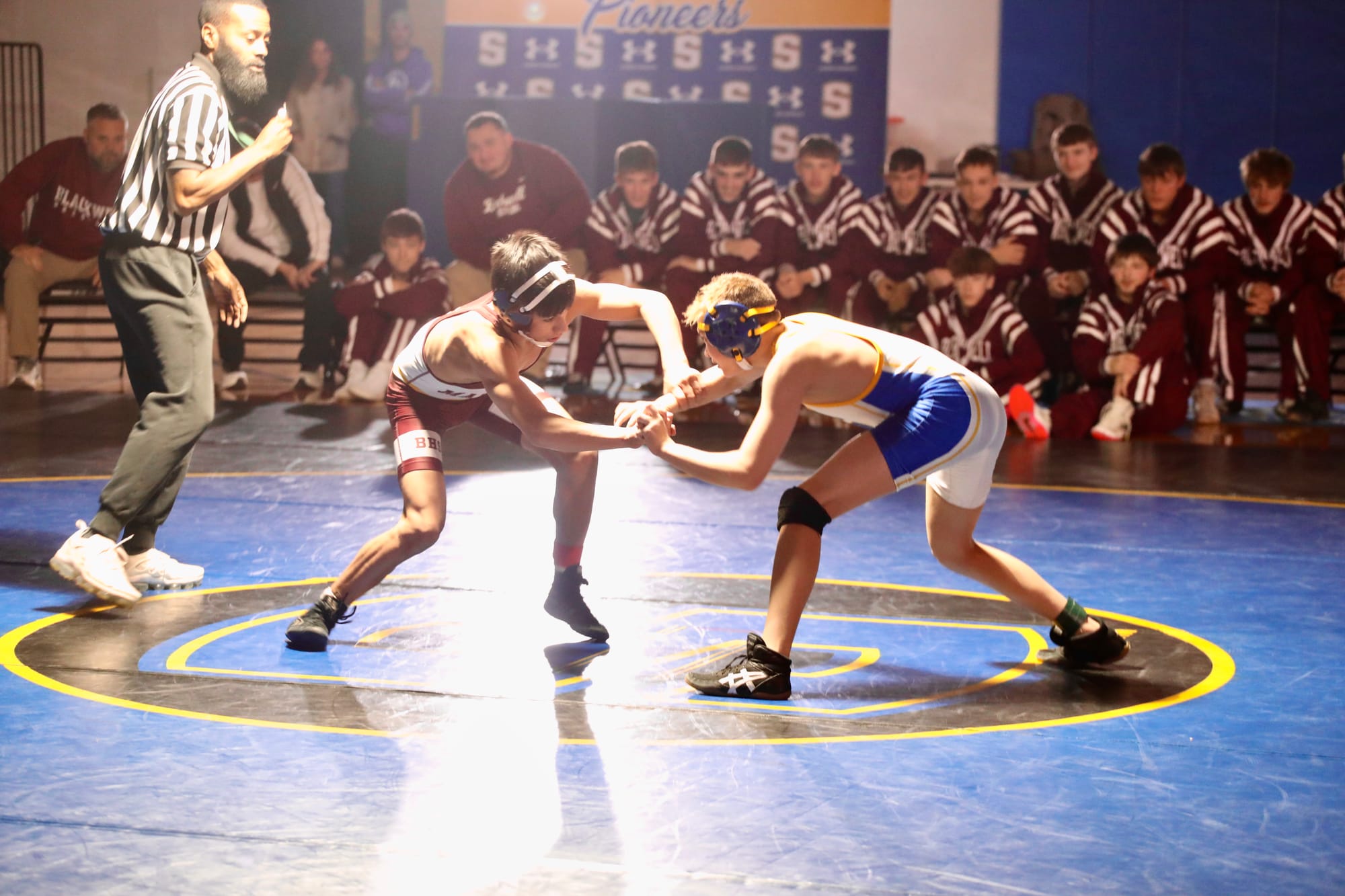 Two teen wrestlers, one in a maroon Blackwell singlet and one in a blue and gold Stillwater singlet, lock hands at the center of a blue mat as a referee looks on. Wrestlers in maroon warmups sit along the edge of the mat in front of a Pioneers banner.