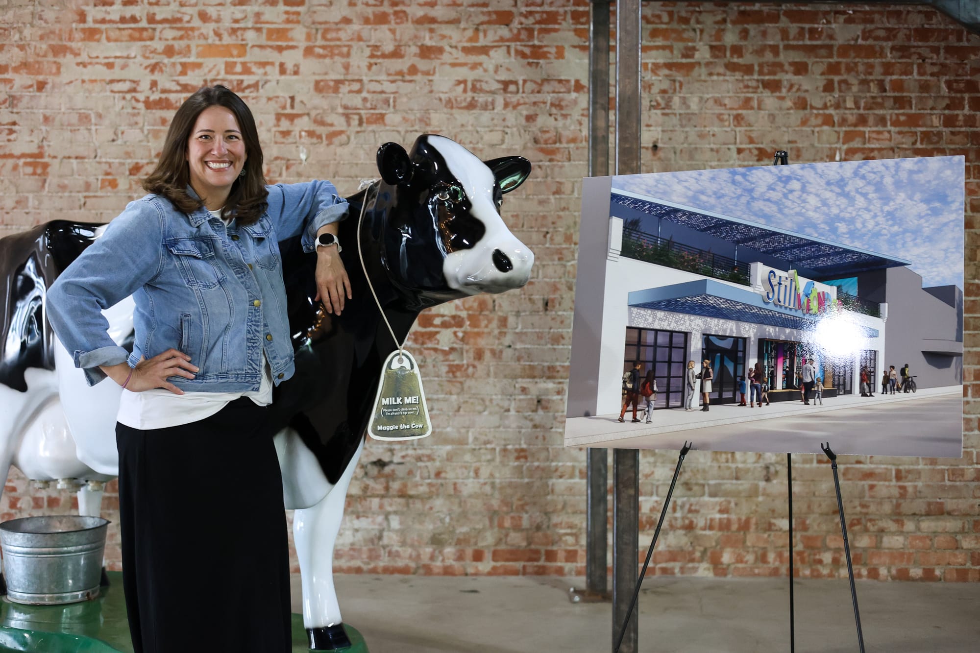 A smiling woman in a denim jacket leans on a black-and-white fiberglass cow beside an easel holding a rendering of the StillWonder children's museum storefront.