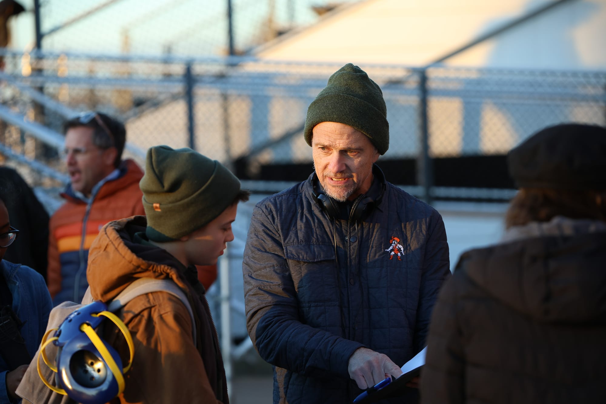 A man in a green beanie and a navy jacket with an OSU Pistol Pete logo points to pages on a clipboard while talking with a teen in a matching green beanie, who carries a blue and gold wrestling headgear on his backpack. The bleachers of a football stadium are behind them.