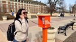 Student in sunglasses and backpack talks on orange OSU phone booth labeled "The Code Calls" on brick campus walkway with students passing in background