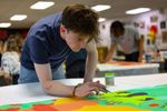 OSU student Jake Warner leans over a large fabric panel, painting with a fine brush in a studio classroom at the Prairie Arts Center, as classmates work in the background.