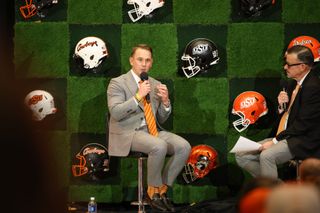 Eric Morris in gray suit with orange tie speaks into microphone during Oklahoma State football coach introduction, with OSU helmets displayed on wall behind him