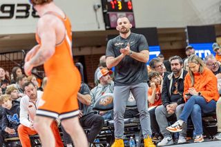 OSU wrestling coach David Taylor stands coaching during a match against Oklahoma at Gallagher-Iba Arena. An OSU wrestler in orange singlet is visible in foreground with fans in background.