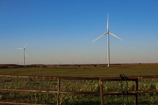 Wind turbines rise above green agricultural fields with a rusty barbed wire fence in the foreground under a clear blue sky in Noble County, Oklahoma