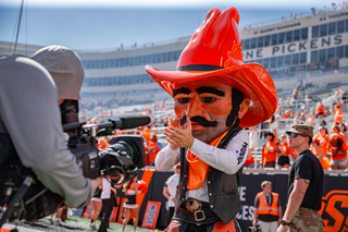 Pistol Pete mascot in orange outfit and cowboy hat stands on sideline at Boone Pickens Stadium with crowd in background during Oklahoma State football game