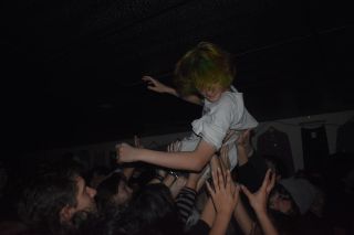 A crowd surfer is held aloft by multiple hands in a dark concert venue with a low black ceiling visible above.