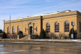 Historic tan brick neoclassical library building with "PUBLIC LIBRARY" text on facade, featuring arched windows, central entrance with pediment, and curved staircases.