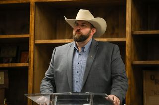 Dillon Travis wearing tan cowboy hat and gray suit jacket stands at clear podium while speaking at indoor campaign event with wooden cabinetry visible in background.