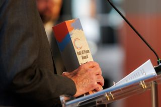 Steve Irby holds a wooden Hall of Fame Award trophy with red, blue, and gray paint strokes at a clear podium during the Stillwater Chamber Awards Gala.
