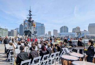 A high school concert band in black uniforms performs on the USS Midway flight deck, surrounded by vintage aircraft and the San Diego skyline.