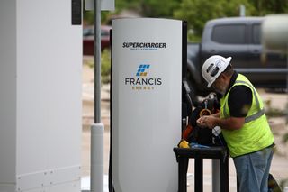 A construction worker in a white hard hat and yellow safety vest works on a white Francis Energy-branded Tesla Supercharger unit in a Stillwater parking lot. 