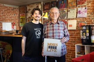 Henry Ramsay and Steve Irby stand together inside Velvet Fudge, holding the Moses Live album, with records displayed on exposed brick walls behind them. 