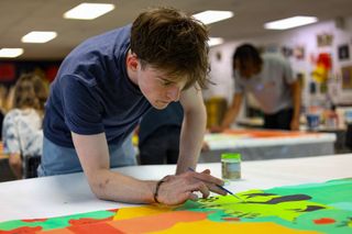 OSU student Jake Warner leans over a large fabric panel, painting with a fine brush in a studio classroom at the Prairie Arts Center, as classmates work in the background.