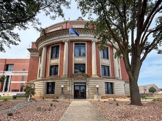 The Payne County Courthouse in Stillwater, Oklahoma, with the American and Oklahoma state flags flying from a flagpole at the entrance and a large tree to the right.