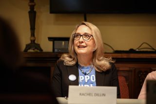 Rachel Dillin, wearing a black blazer and campaign button, sits at a table during a candidate forum. A nameplate reading "Rachel Dillin" is visible in the foreground.