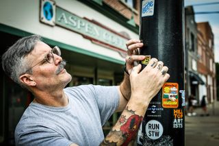 Patrick Jalbert places a Stillwegian sticker on a downtown Stillwater pole outside Aspen Coffee, alongside stickers from Mid South and other local organizations.